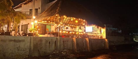 Un establecimiento frente a la playa con una estructura de techo de paja, mesas al aire libre y una pantalla de televisión visible.