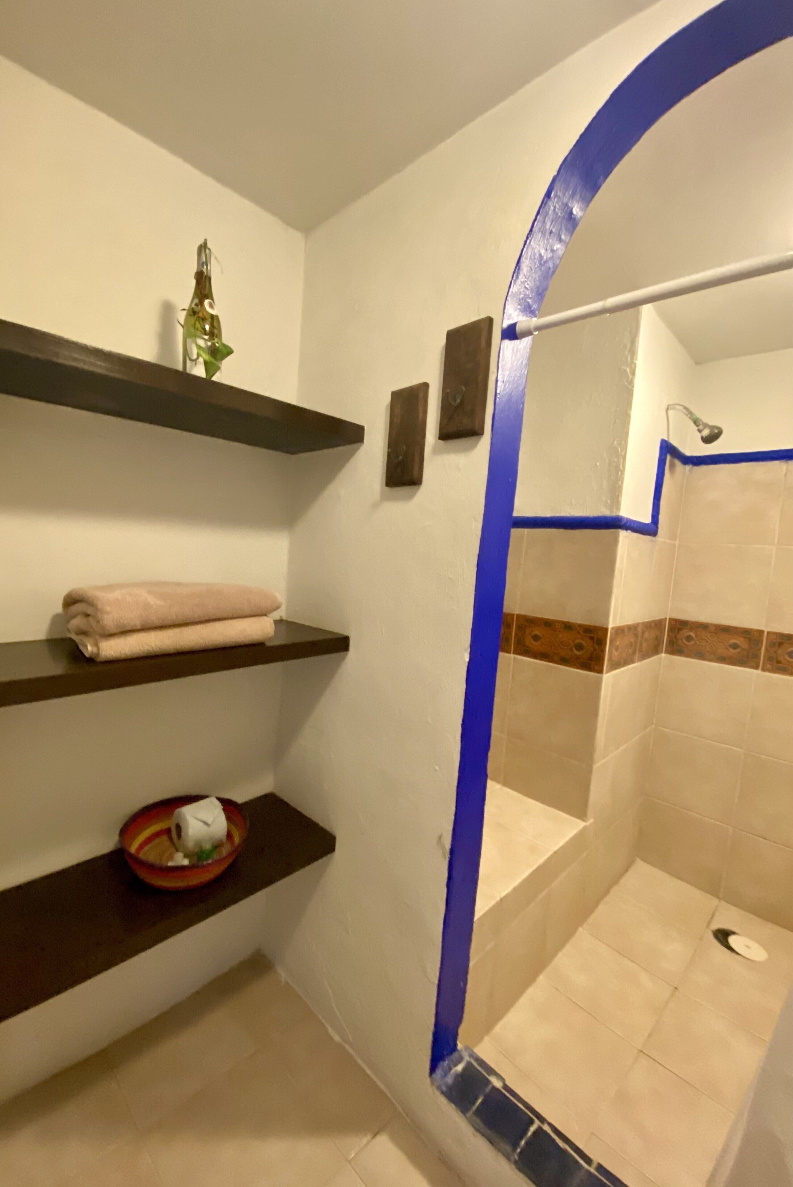 Un baño con ducha, azulejos color beige, una pared de acento azul y estantes con toallas y artículos de aseo.