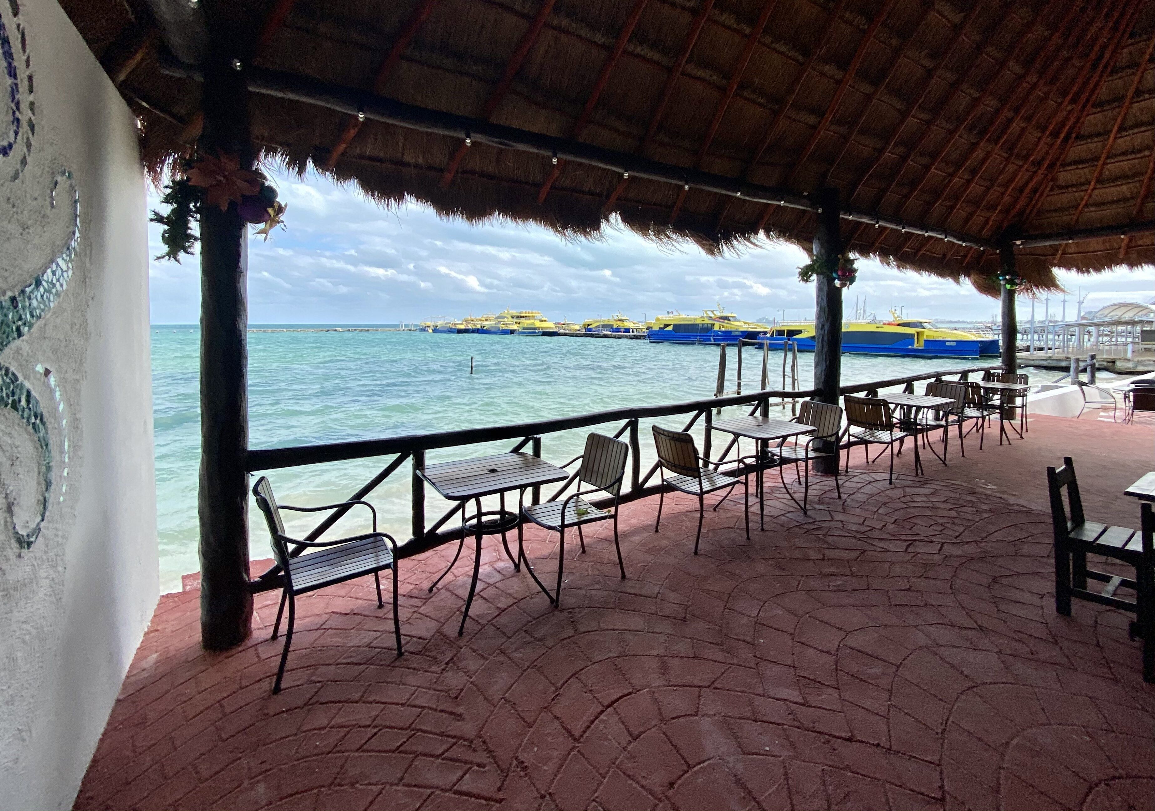 A covered outdoor seating area with tables and chairs, overlooking a body of water with boats.