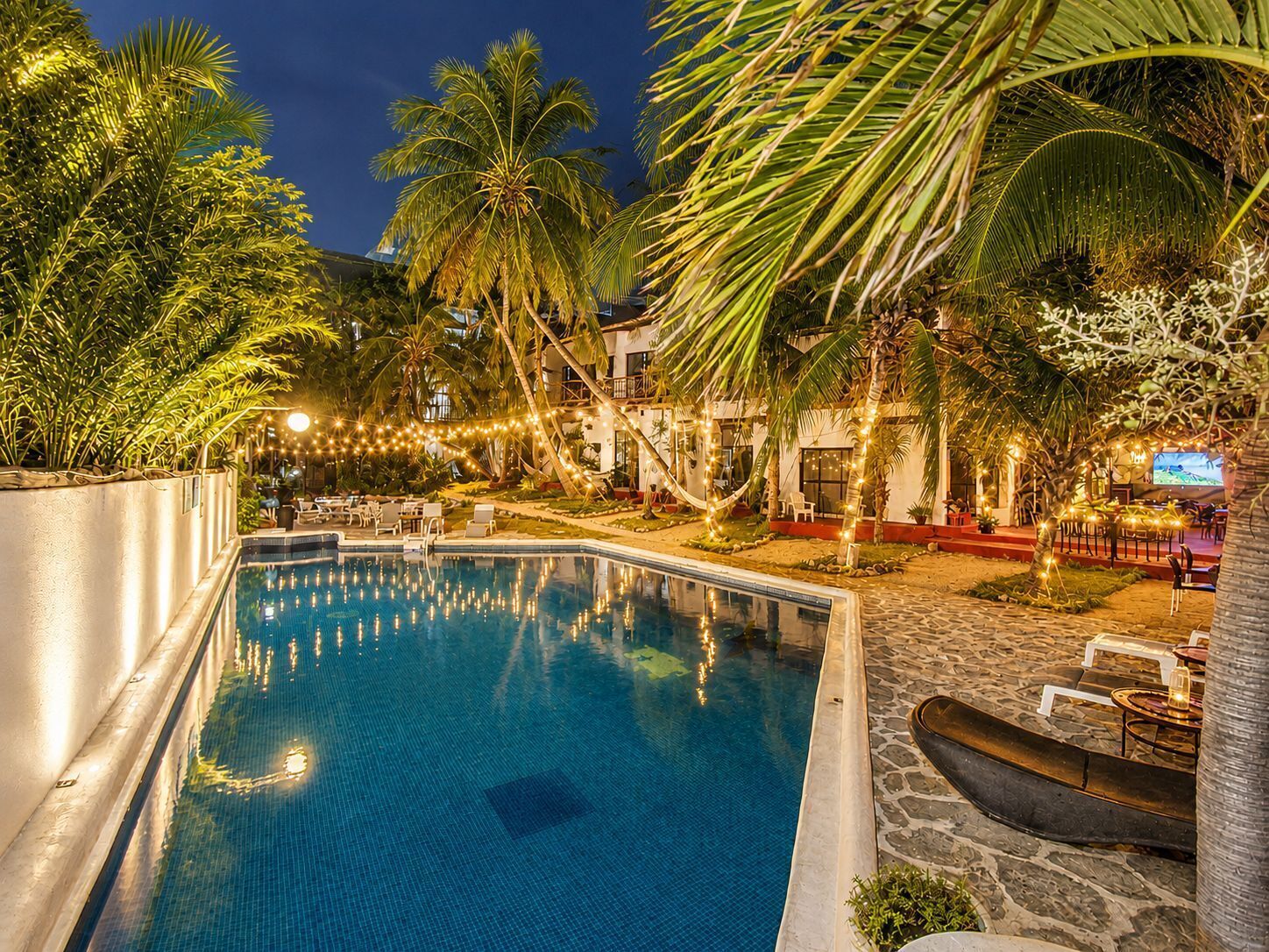 A swimming pool surrounded by palm trees and outdoor seating, with a building in the background.