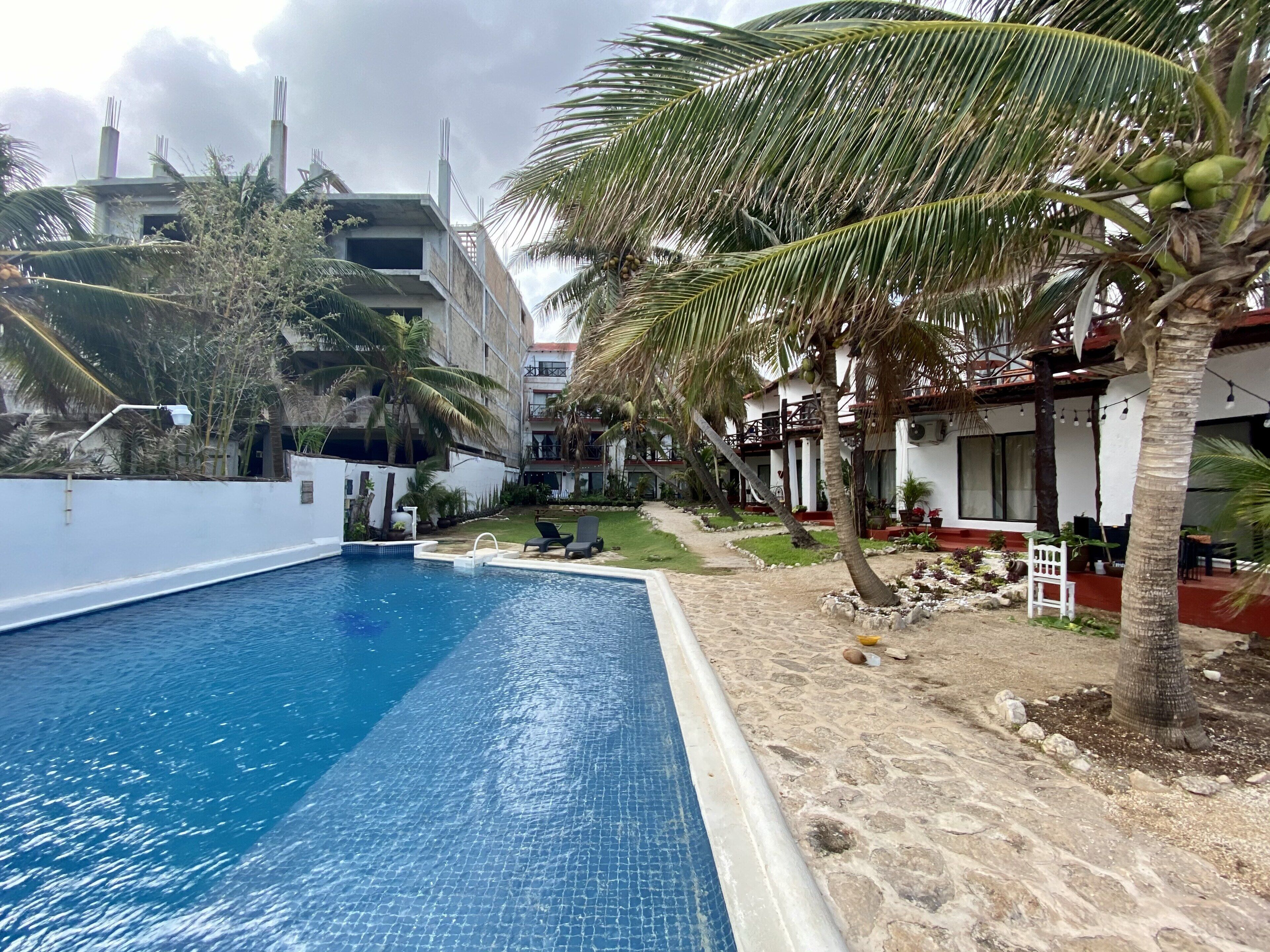 Une piscine dotée d’une terrasse carrelée, entourée de palmiers et bordée d’un bâtiment avec un balcon.