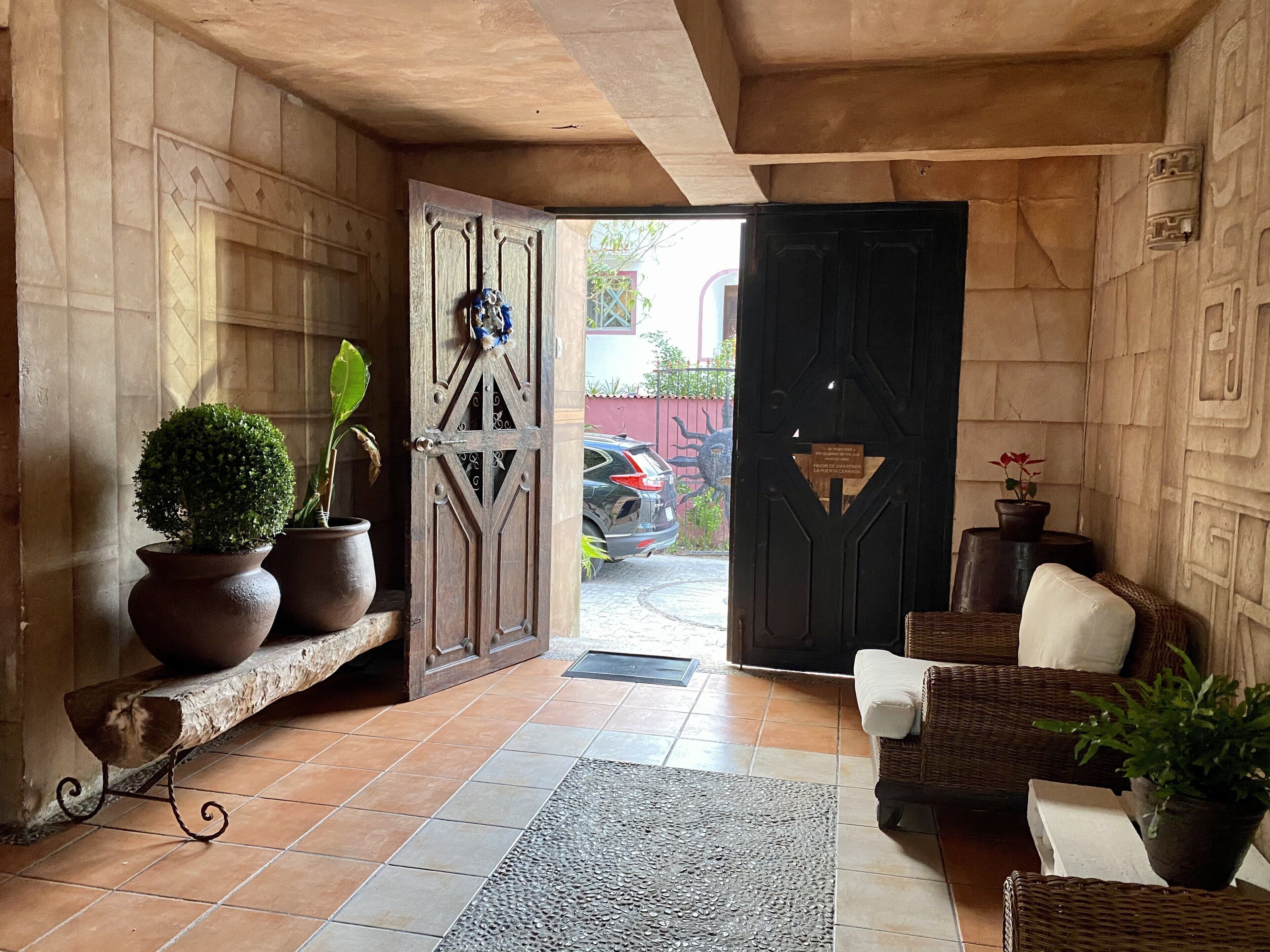 Un interior de madera con suelo de baldosas, una puerta que da al exterior, un sofá de mimbre y plantas en macetas.