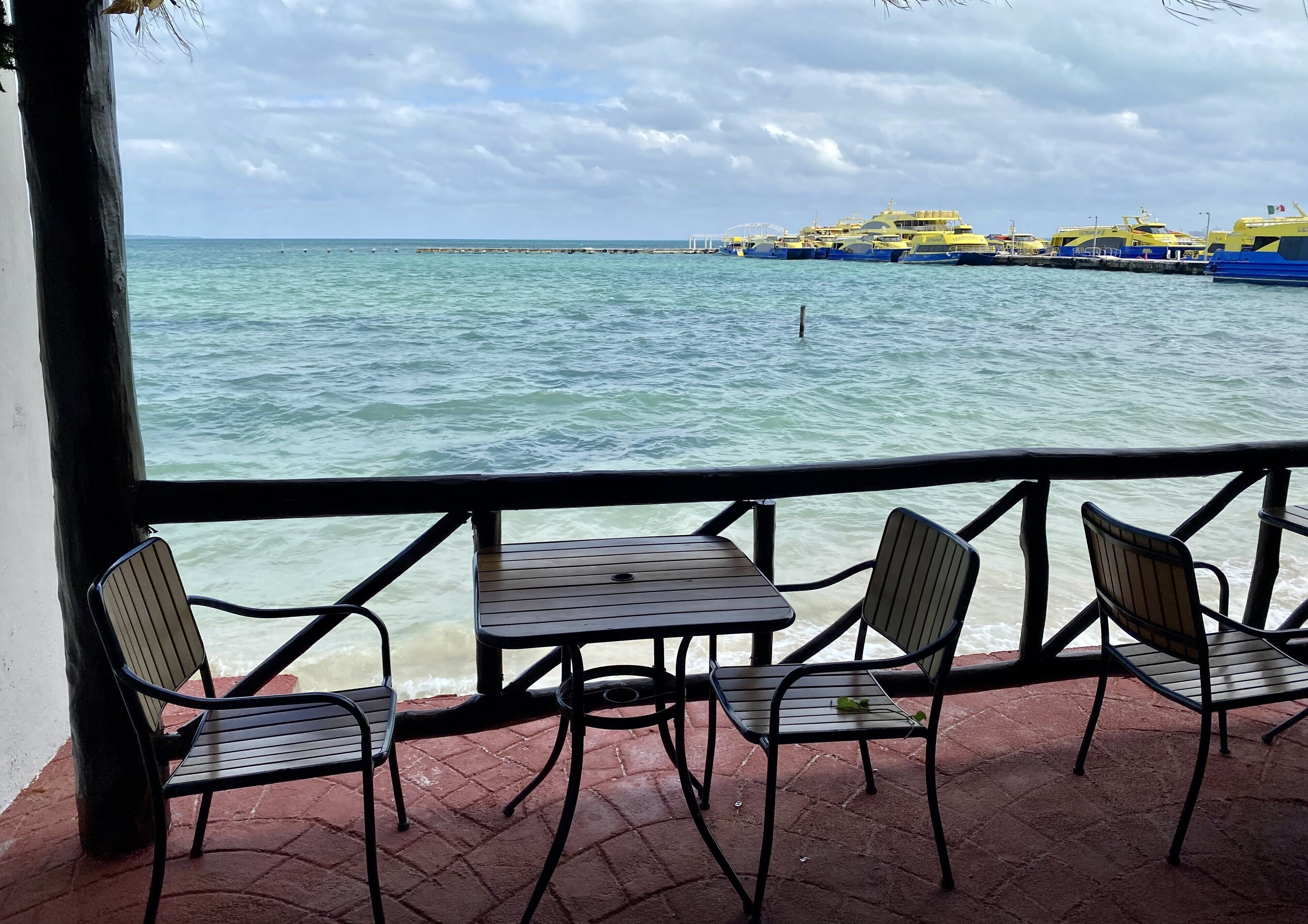 A seaside patio with a table and chairs, overlooking the sea and docked boats.