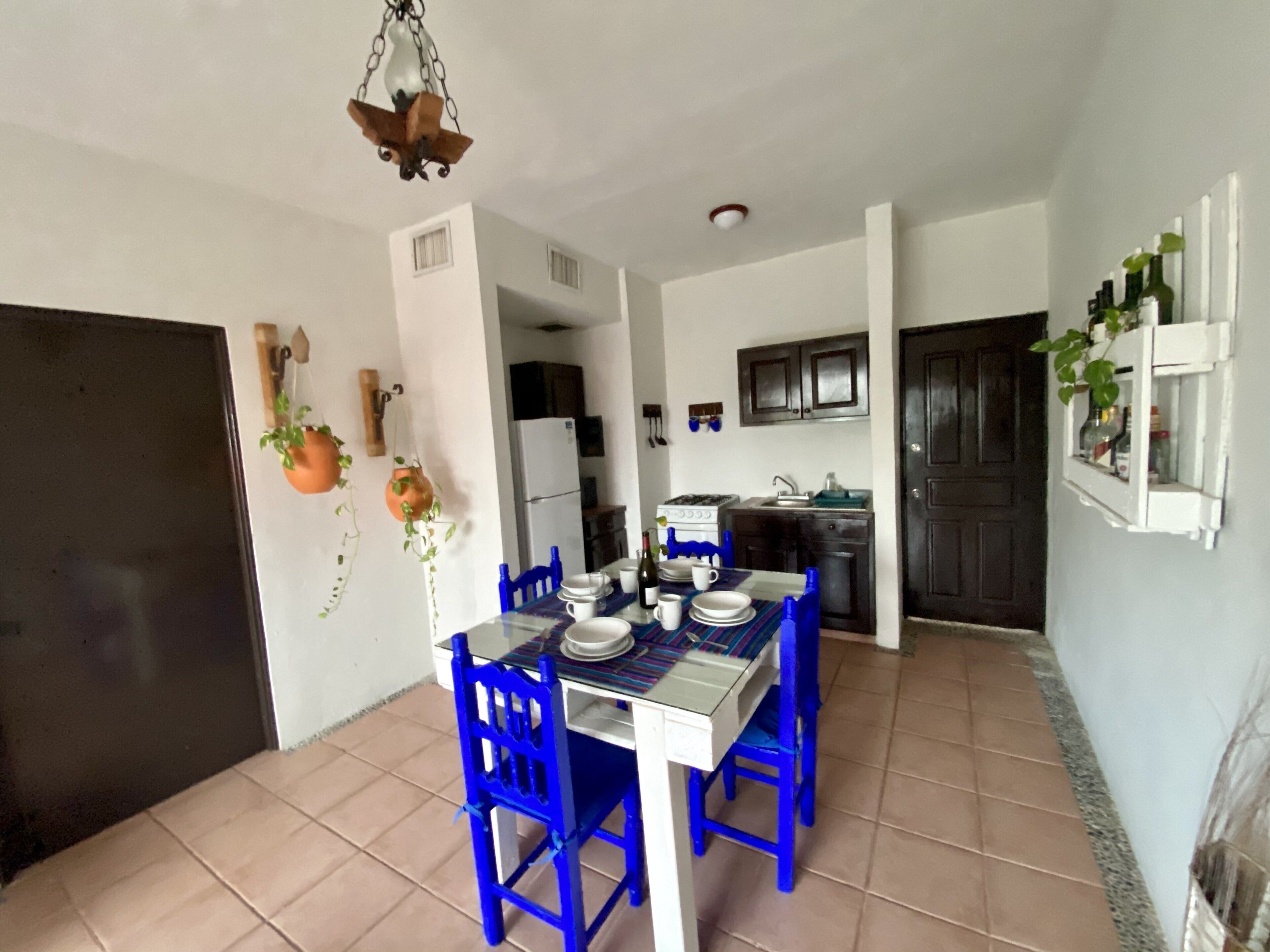A dining area with a table set for four, a kitchen in the background, and a door leading to another room.