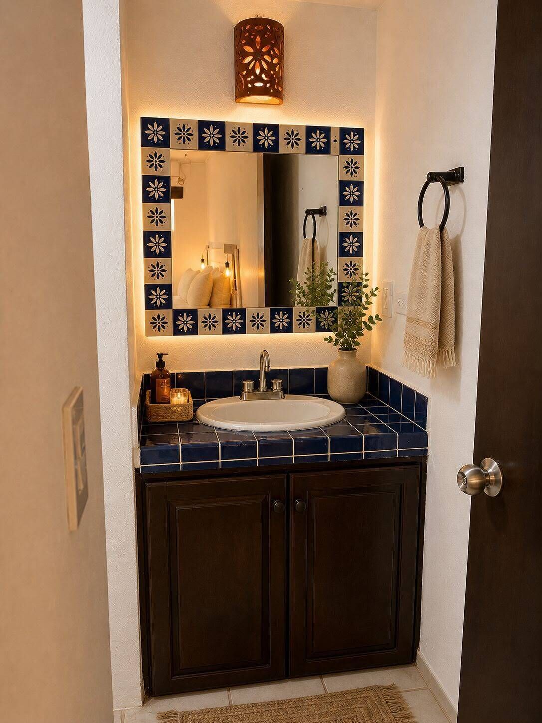 Un baño con un mueble de madera oscura, un lavabo blanco y un espejo con un borde de azulejos decorativos.