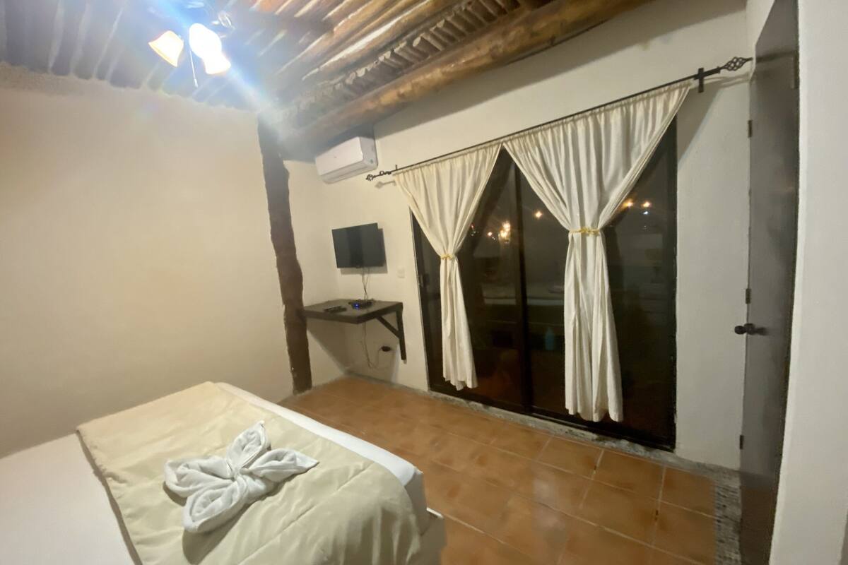 Ein Schlafzimmer mit einem Bett, einem Fernseher, einem Fenster mit Vorhängen und einem Deckenventilator.