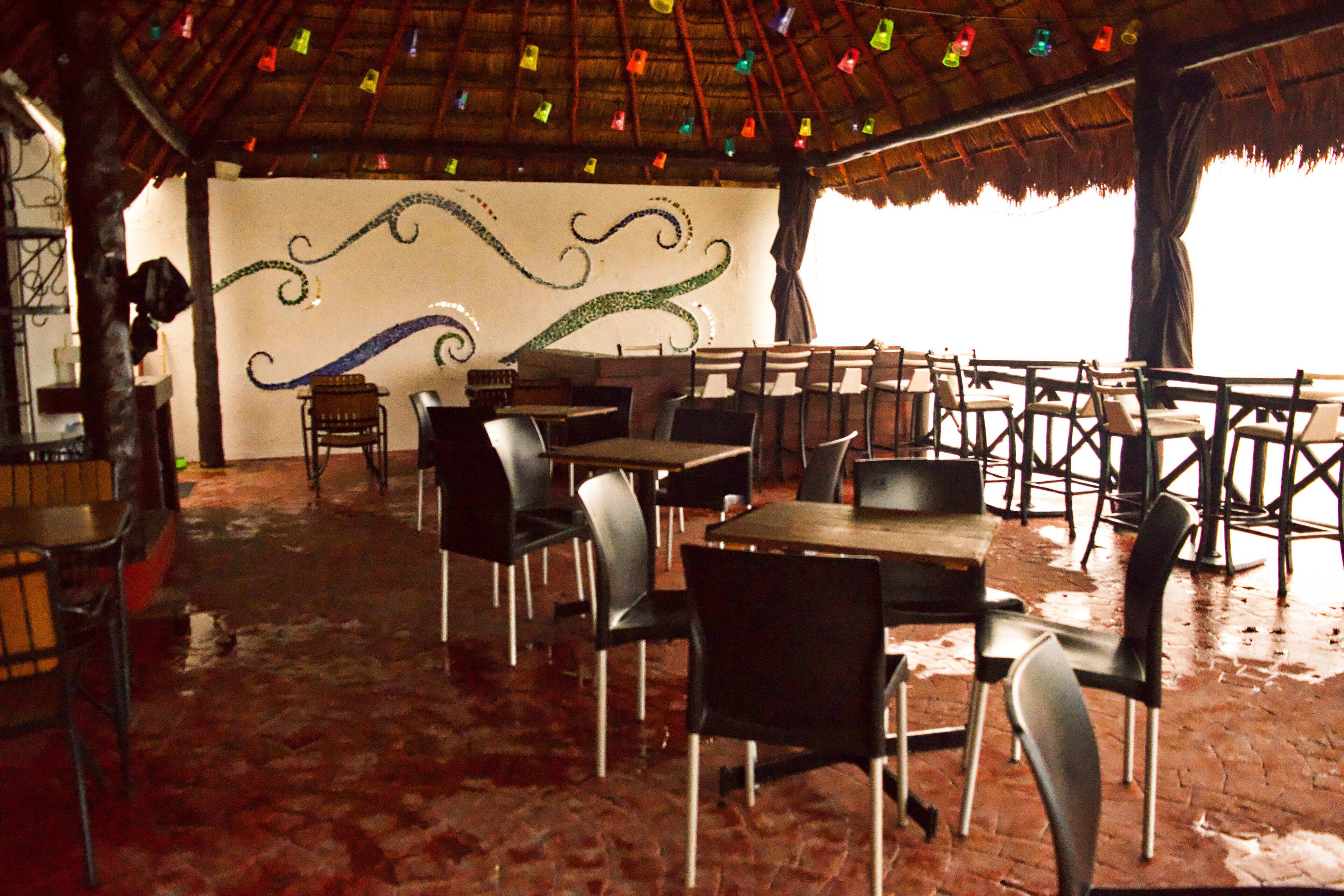 An outdoor dining area with tables and chairs, a thatched roof, and a mural on the wall.