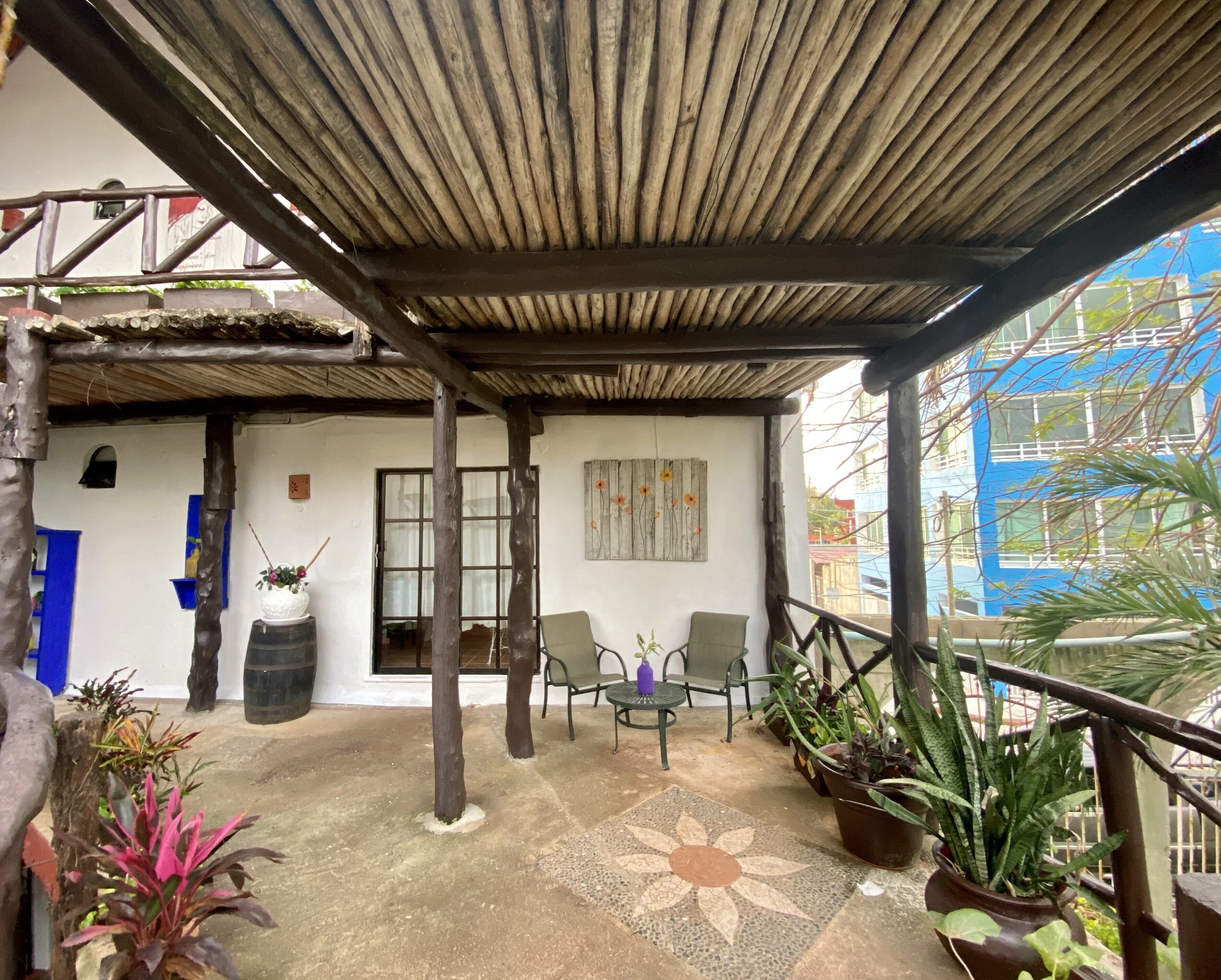 A covered outdoor area with a concrete floor, two chairs, potted plants, and a wooden ceiling structure.