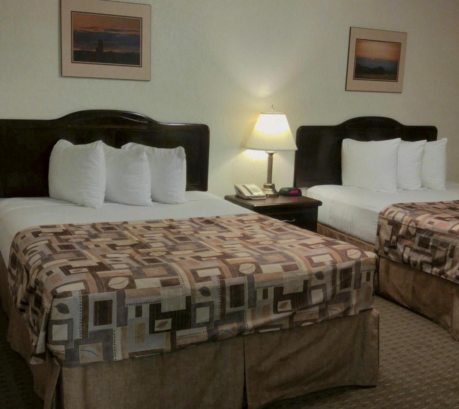 Habitación de hotel con dos camas, una mesita de noche con lámpara y dos cuadros enmarcados en la pared.