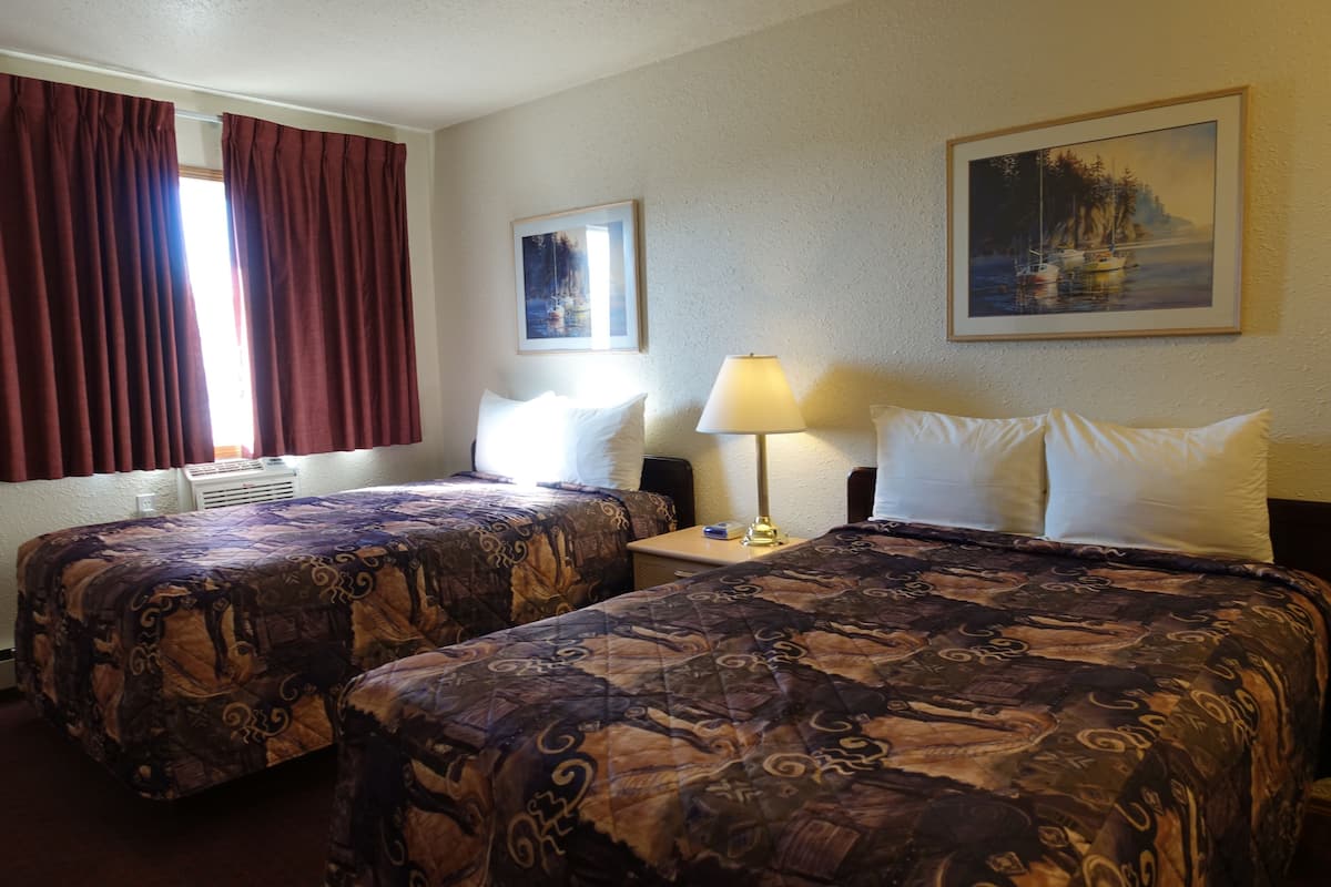 Quarto de hotel com duas camas, uma mesinha de cabeceira, um abajur e duas pinturas nas paredes.