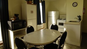 Een kleine keuken met een eettafel, stoelen, een magnetron, een koelkast, een fornuis en een klok aan de muur.