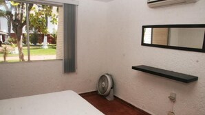 A room with a bed, a standing fan, a window with a view of a garden, and a wall-mounted shelf. - Casa Moises (Mazatlán)
