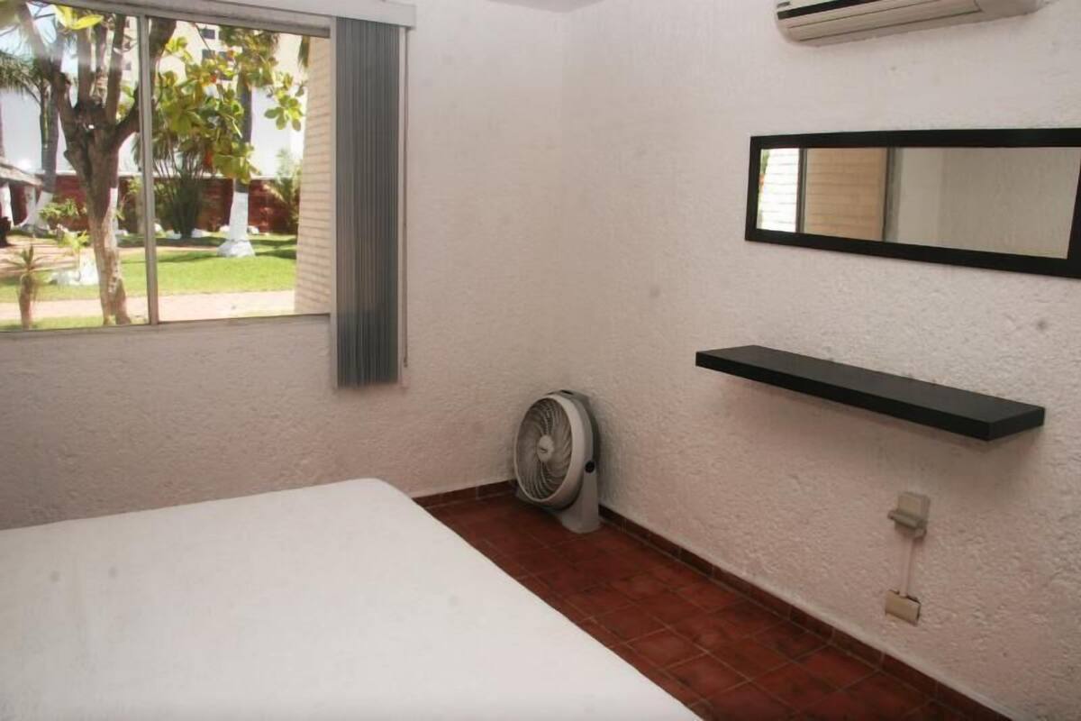 Una habitación con cama, ventilador de pie, ventana con vista a un jardín y un estante en la pared.