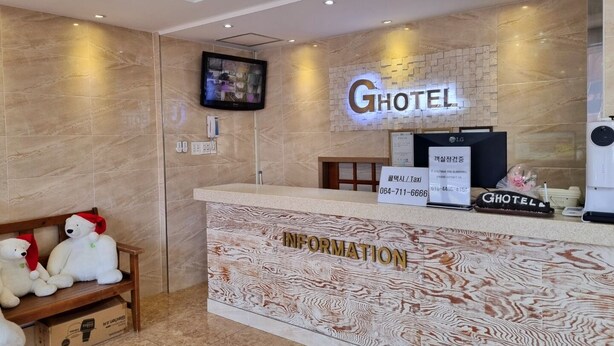 A hotel reception area with a marble wall, a mounted television, and a reception desk labeled "HOTEL" and "INFORMATION". - Hotel G (Jeju City)