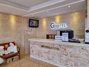 A hotel reception area with a marble wall, a mounted television, and a reception desk labeled "HOTEL" and "INFORMATION". - Hotel G (Jeju City)