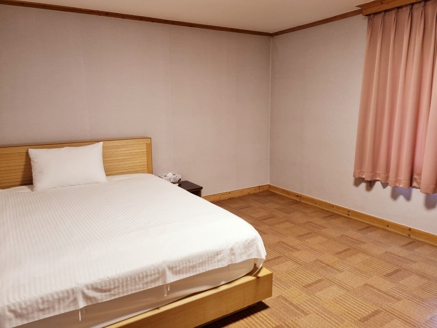 A neatly made bed in a corner room with a wooden headboard, a small bedside table, and a pink curtain on the window.