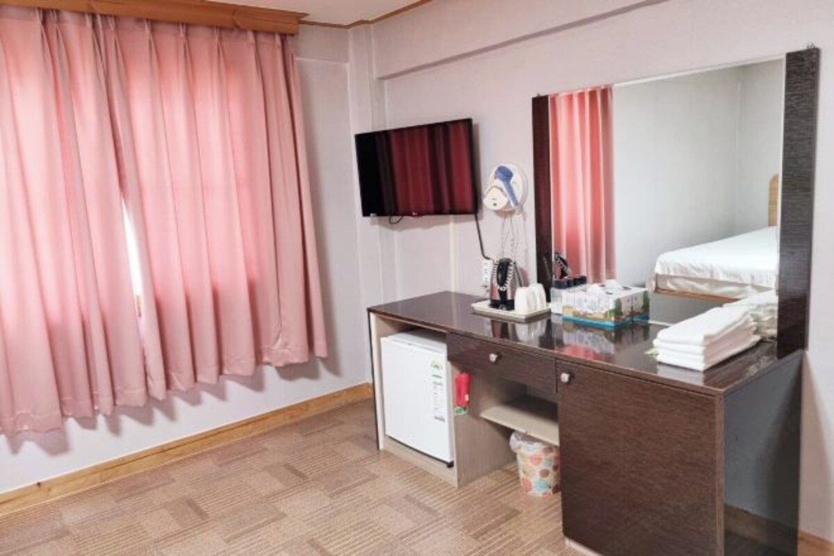 A hotel room with a bed, a desk with a mirror, a television, a small refrigerator, and pink curtains.