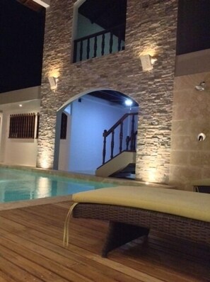 A pool area with a wooden deck and a stone wall. There is a staircase leading to an upper level.