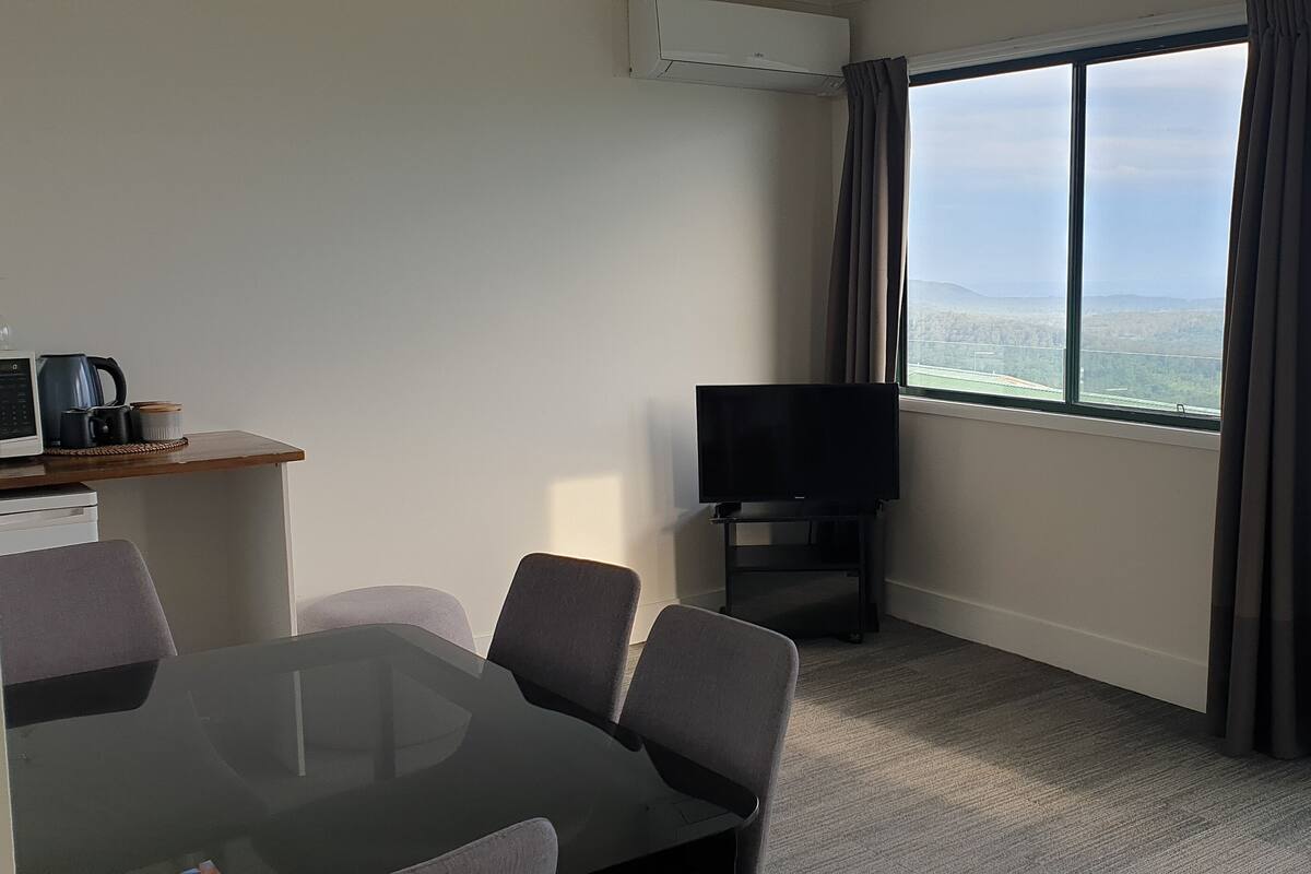 Ein modernes Hotelzimmer mit großem Fenster, Flachbildfernseher, Glastisch und Sitzecke.