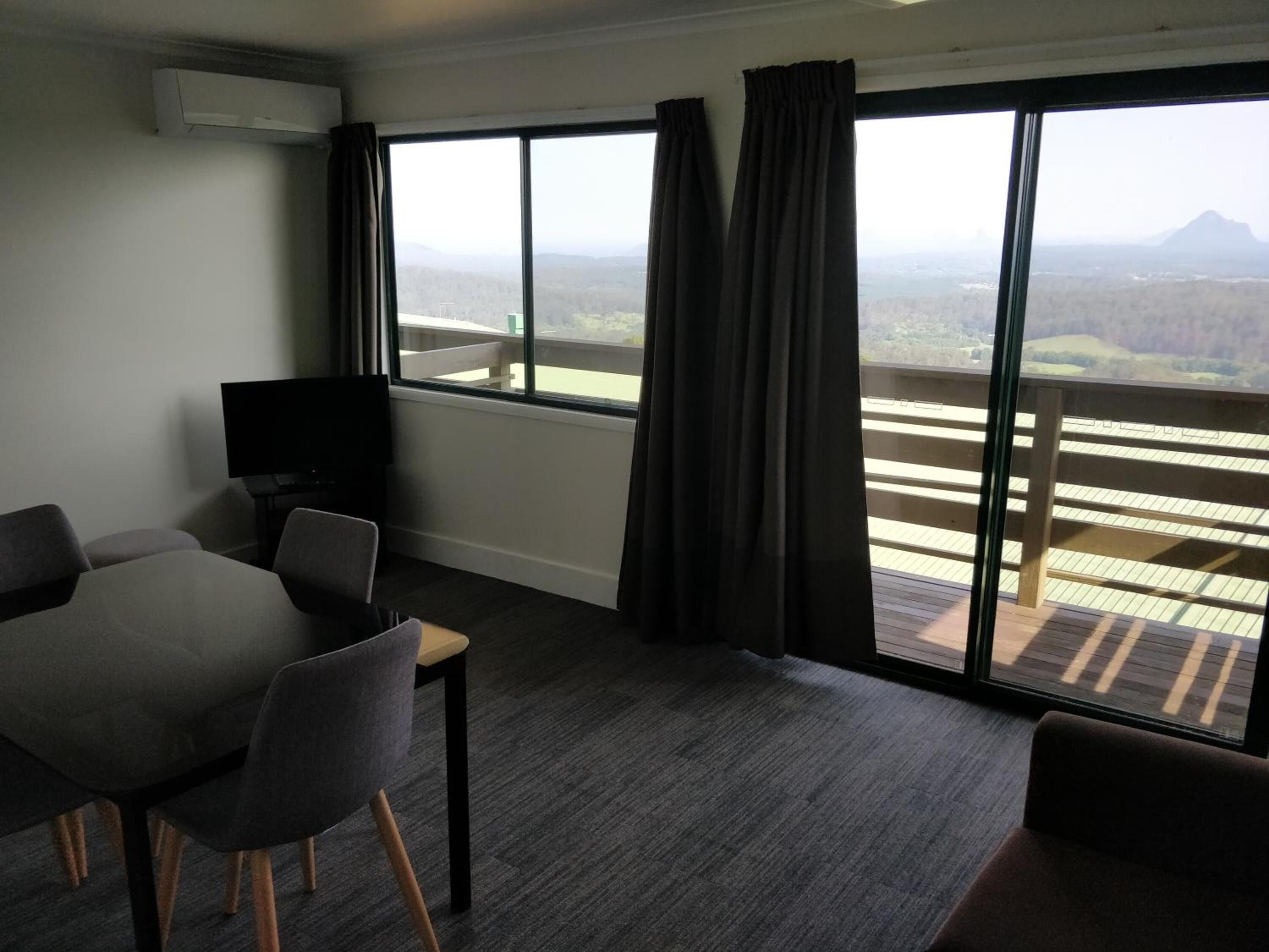 A hotel room with a balcony, a dining table with chairs, a flat-screen TV, and a large window with curtains.