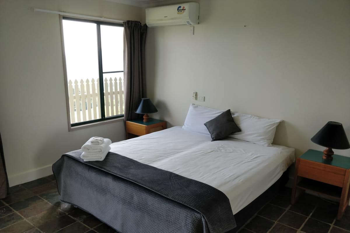 Ein Hotelzimmer mit einem Bett, zwei Nachttischen, einem fensterbelaubten Fenster, einer Klimaanlage und einem gefliesten Boden.