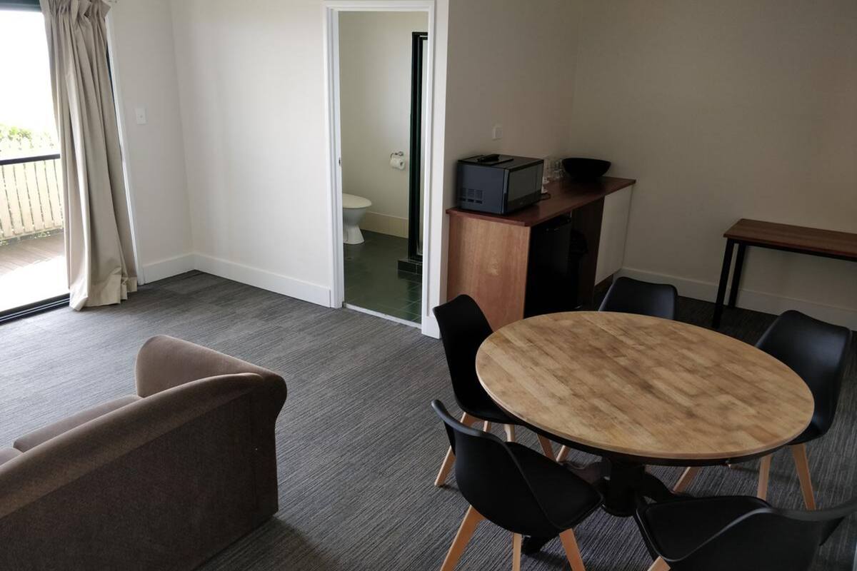 Ein Zimmer mit einem runden Holztisch und vier schwarzen Stühlen, einem braunen Sofa, einem kleinen Schrank mit Fernseher sowie einem Blick auf einen Balkon.