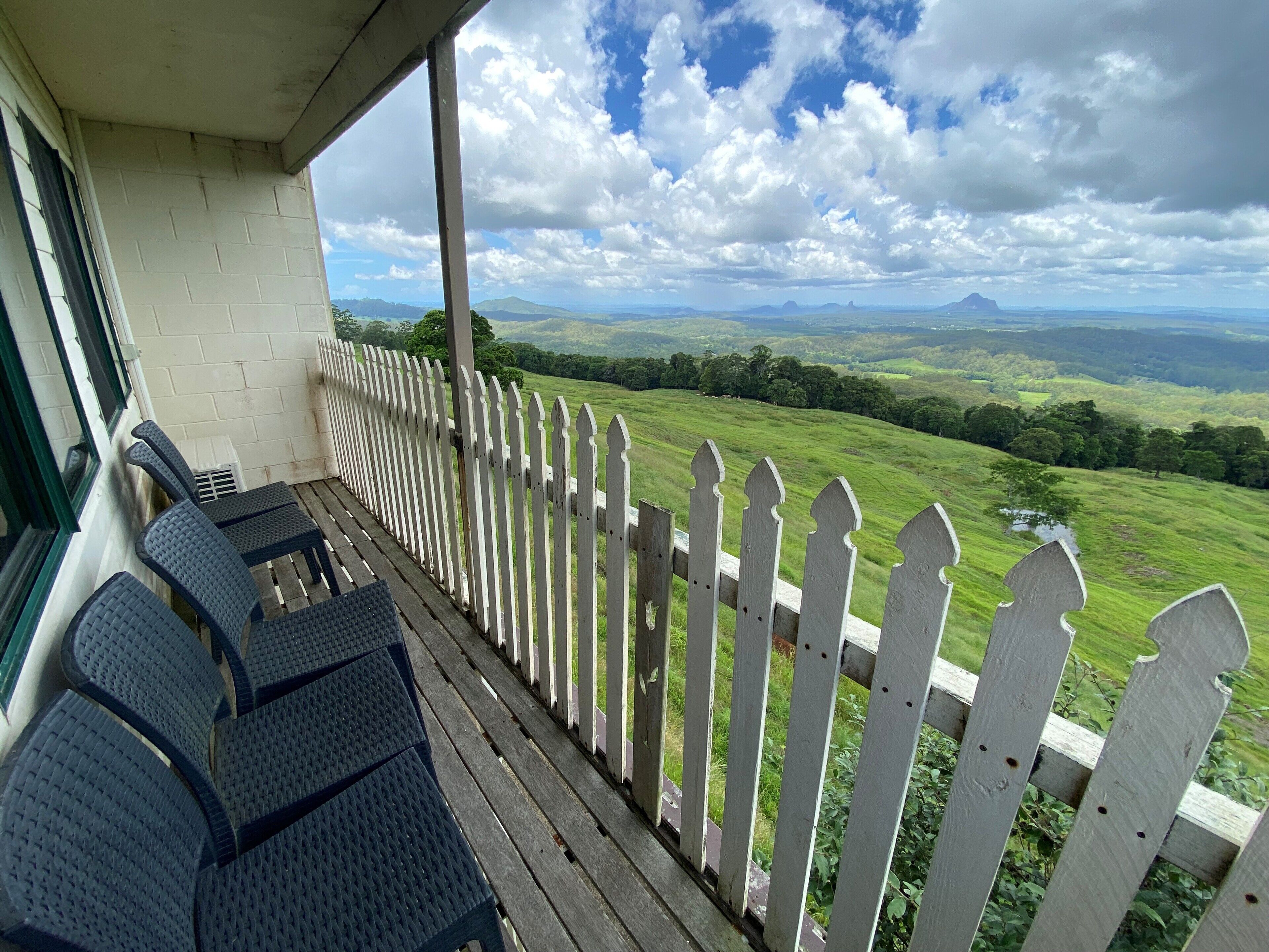 A covered outdoor area with a white picket fence, a table with chairs, and a view of green hills and distant mountains.