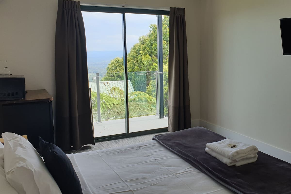 Ein Hotelzimmer mit Bett, Flachbildfernseher, Fenster mit Vorhängen und Blick auf Grünfläche.