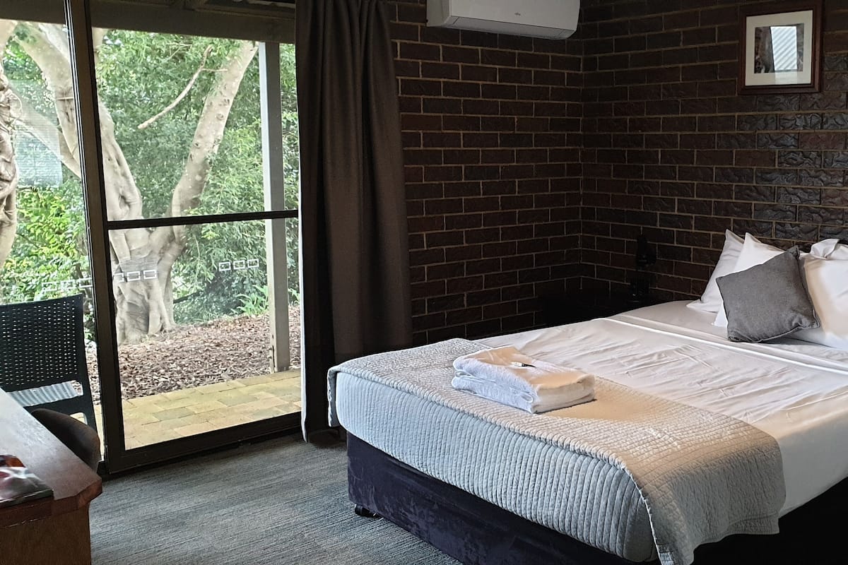 Ein Schlafzimmer mit einem großen Bett, einem Nachttisch, einem Fenster mit Blick auf Bäume und einer Ziegelwand.