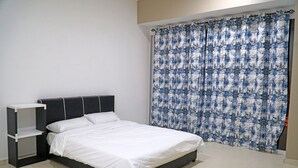A neatly made bed with white bedding, a dark headboard, and a small white nightstand with a shelf. A patterned curtain covers the window. - CVR Homestay Melaka (Jasin)