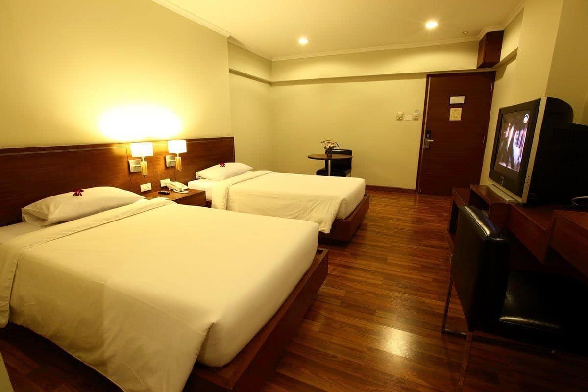 A hotel room with two beds, a desk, a TV, and a small table with a vase.
