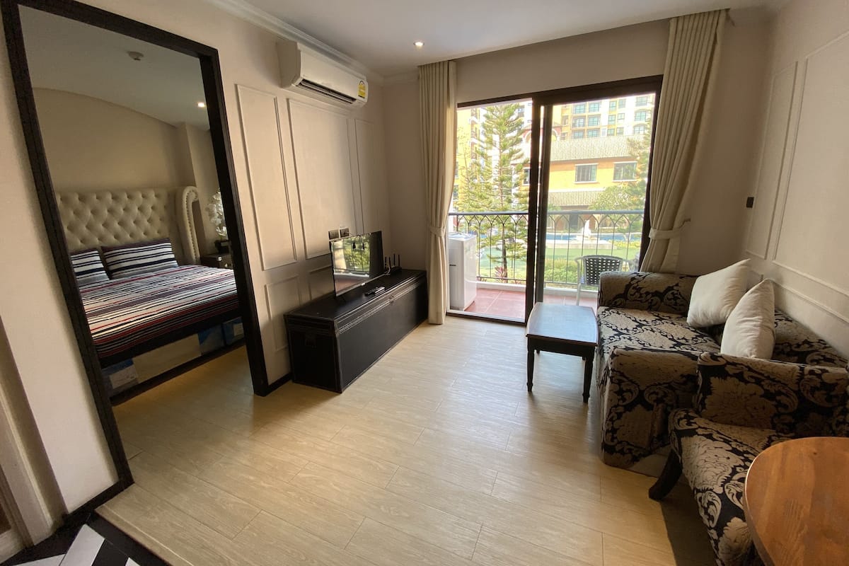 Ein Hotelzimmer mit Bett, einer Sitzecke, einem Fernseher, einem kleinen Tisch und einem Balkon mit Blick auf Gebäude und Bäume.