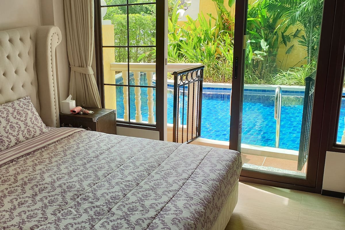 Ein Schlafzimmer mit einem großen Bett, einem Nachttisch und Blick auf einen Swimmingpool durch eine Glastür.