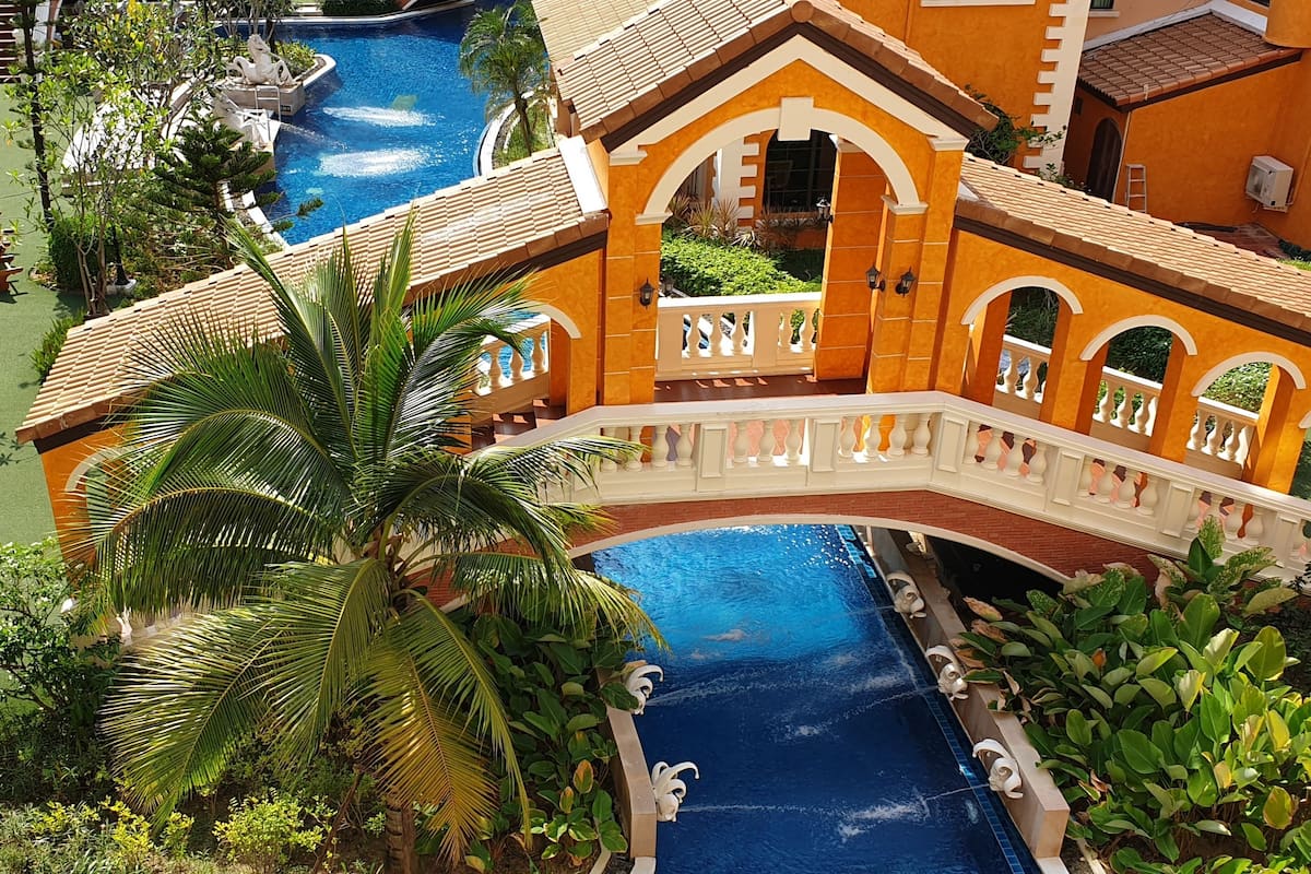 Ein Resort mit mehreren Schwimmbecken, einer zentralen Brücke und üppiger Vegetation.