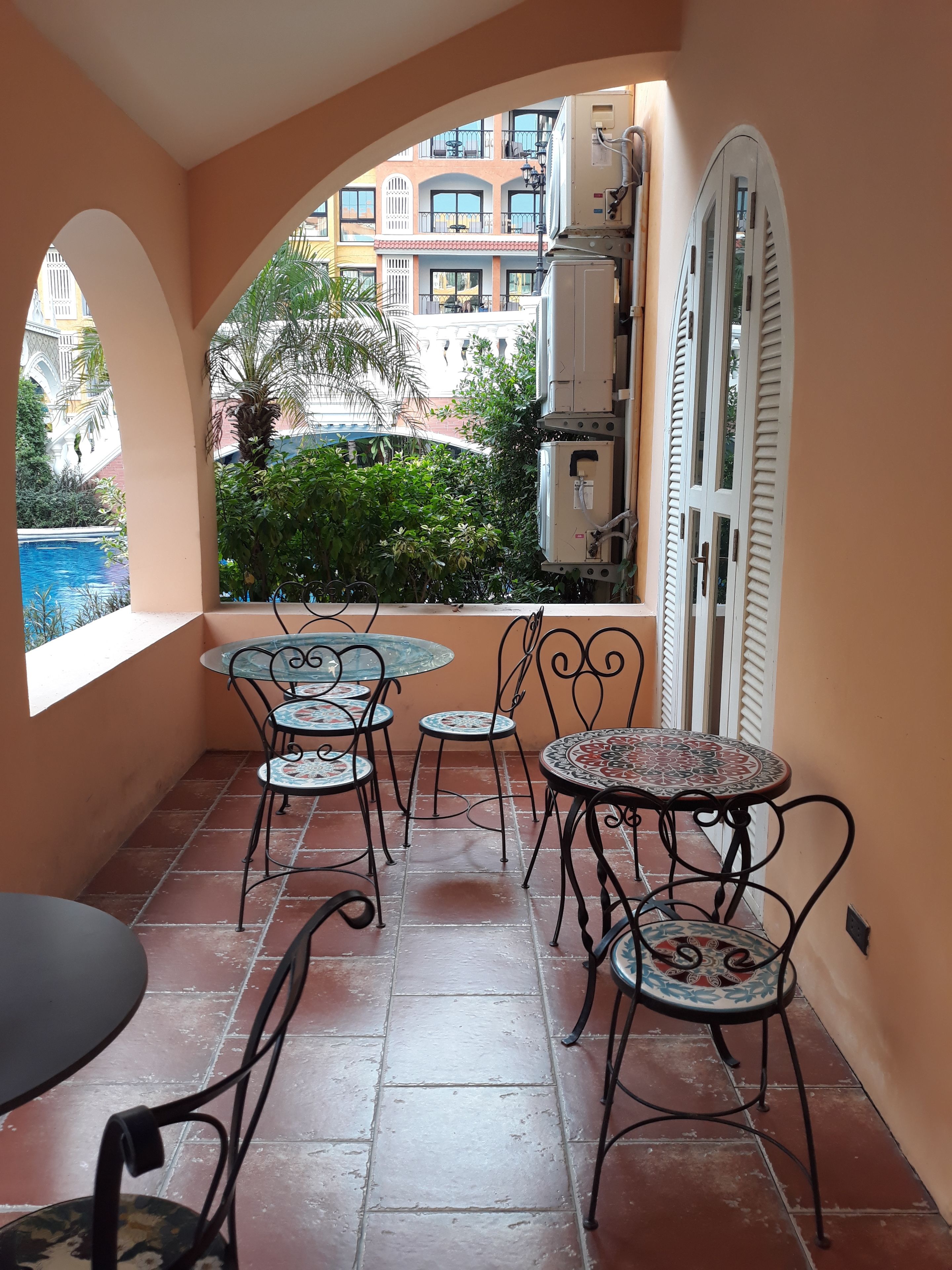 A small patio with a pool view, featuring a table and chairs, and a window overlooking a building with balconies.