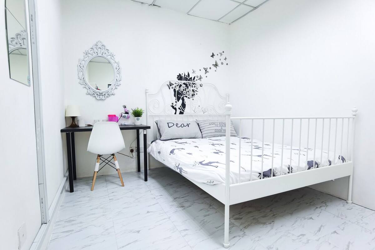 Une chambre à coucher blanche, avec un lit, un bureau, une chaise, un miroir et un motif décoratif sur le mur.