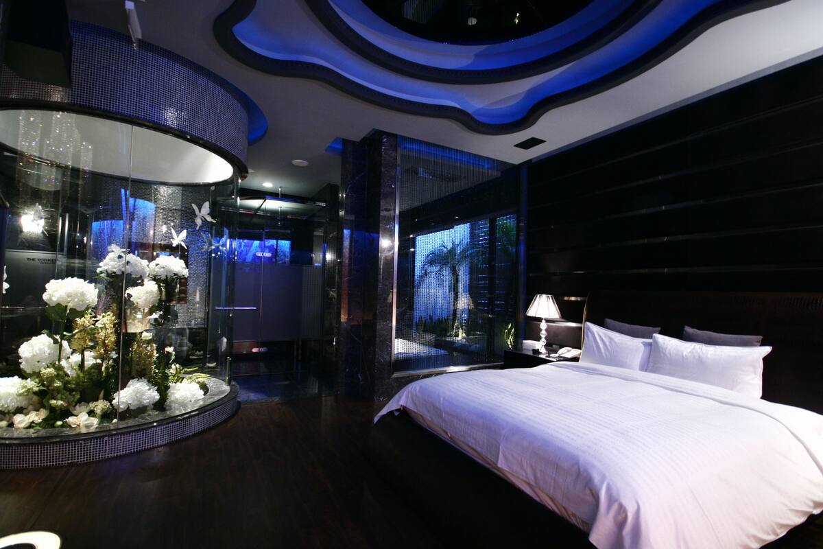 A modern hotel room with a large bed, a glass display with flowers, and a cityscape view.