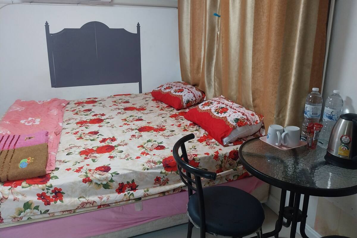 Una cama bien tendida con ropa de cama floral, una sola silla, una mesita con vasos y botellas de agua, y un aire acondicionado de pared.