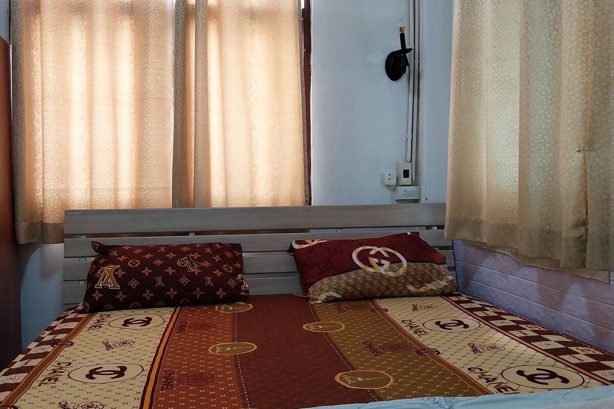 Un dormitorio con cama, cortinas, equipo de aire acondicionado y una lámpara fijada a la pared.