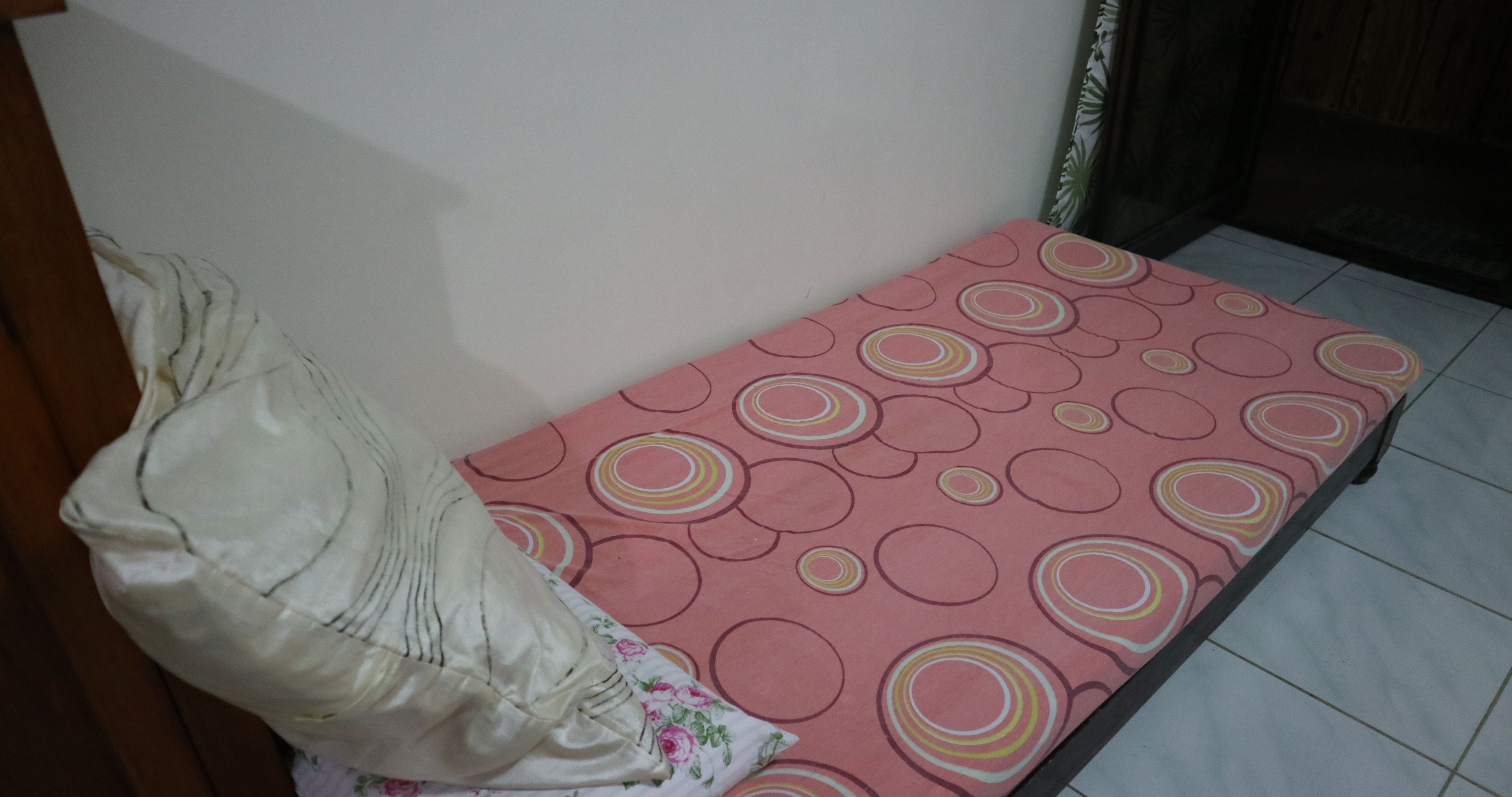 A small, single-bed room with a pink mattress featuring circular patterns, a white pillow, and a floral pillow on the bed.