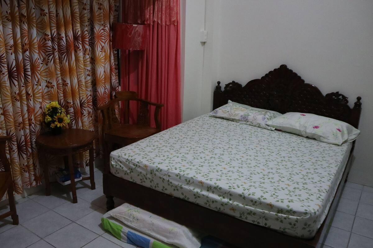 A bedroom with a bed, two chairs, a small table with a vase of flowers, and a curtain with a floral pattern.