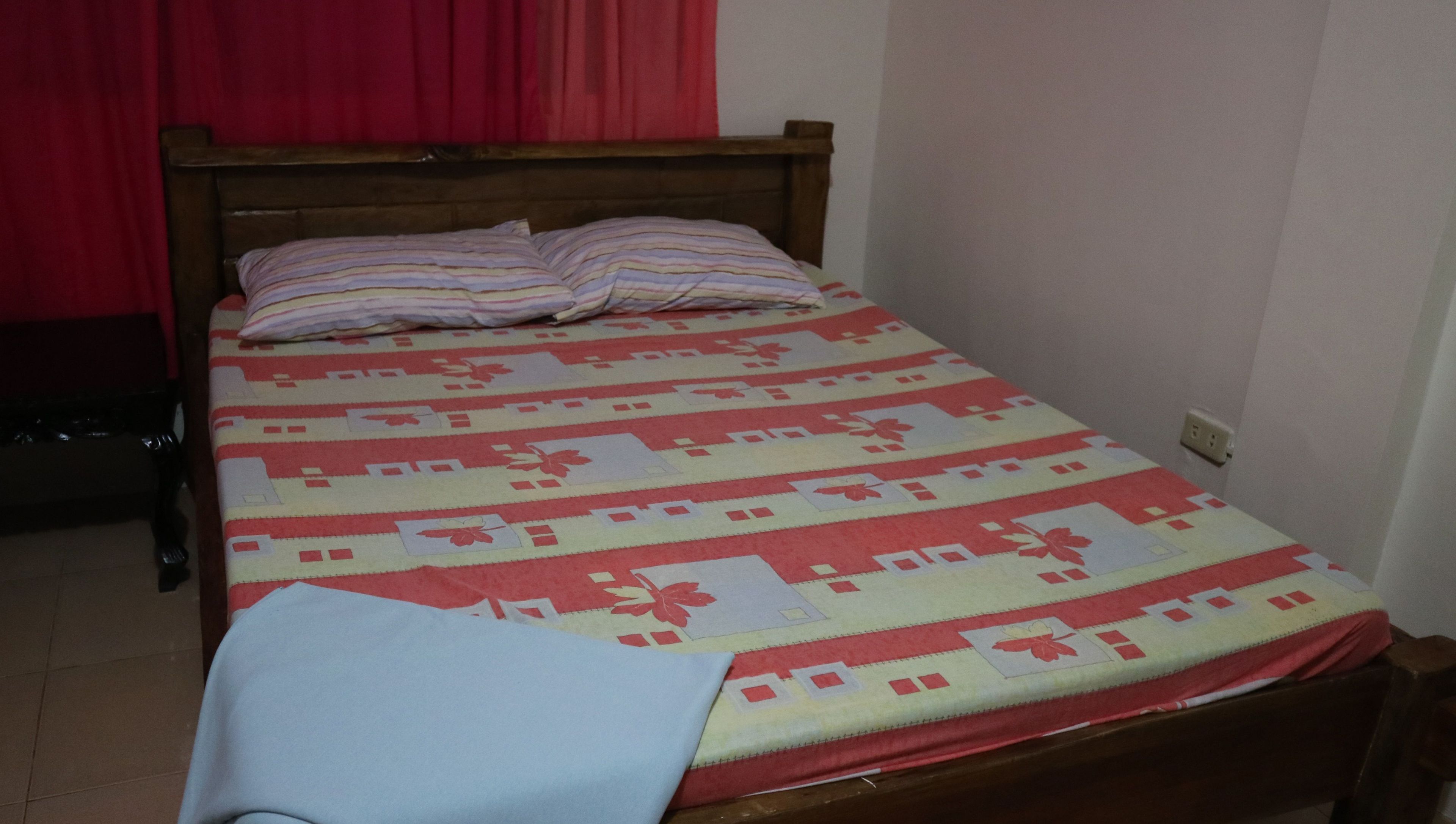 Un lit simple avec une couvre-lit à rayures et des oreillers, une tête de lit en bois et un rideau rouge en arrière-plan.