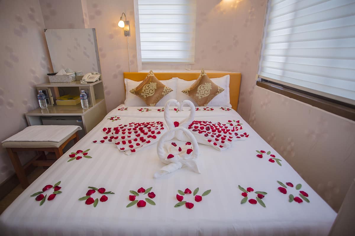 Una cama cuidadosamente arreglada adornada con pétalos de rosa y decoraciones con forma de cisne, una mesita de noche con un teléfono y botellas de agua, y una ventana con persianas.
