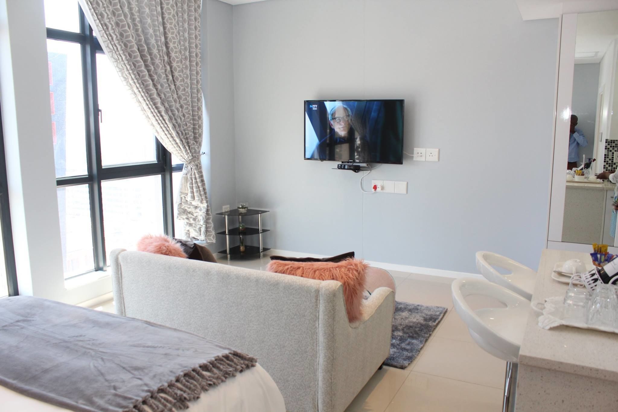 Habitación de hotel moderna con sofá gris, televisor de pantalla plana en la pared, una mesita auxiliar y un baño con espejo y lavamanos.