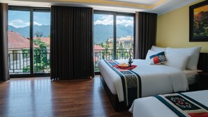 Habitació d'hotel amb un llit gran, un altre més petit, i un balcó que fa vedes a muntanyes i edificis.