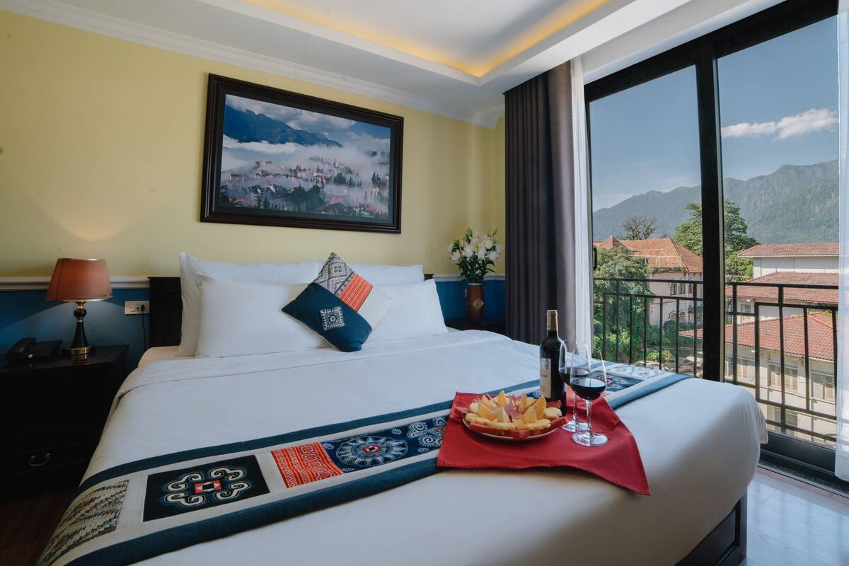A hotel room with a large bed, a painting of a mountain landscape, a nightstand with a lamp, and a view of buildings through a balcony.