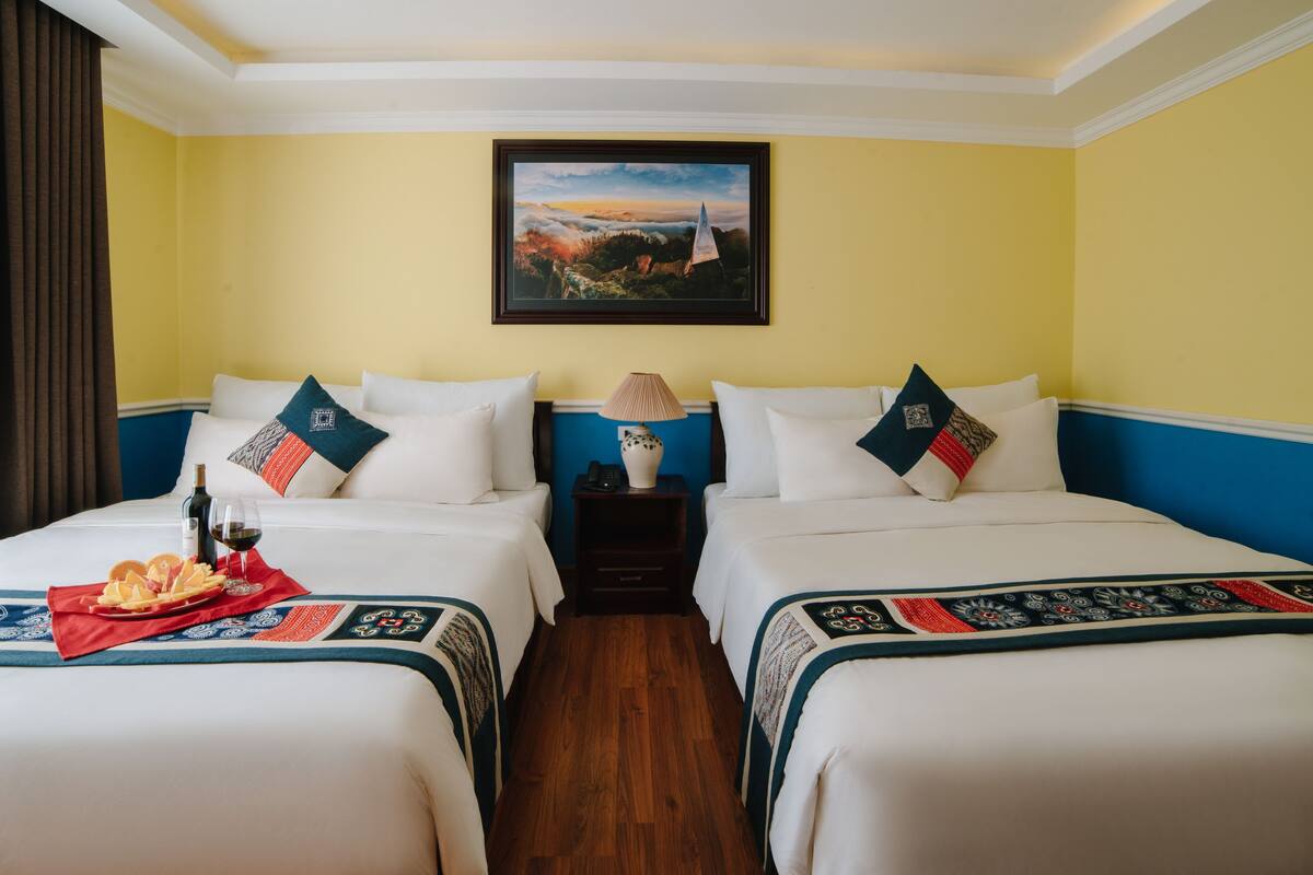 A hotel room with two beds, a painting on the wall, a lamp, and a tray with food and drink on the beds.