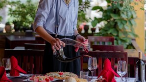 Una persona atent als dinàrgants a un restaurant a l'aire lliure, amb llum decorativa penjant i verdura al fons.