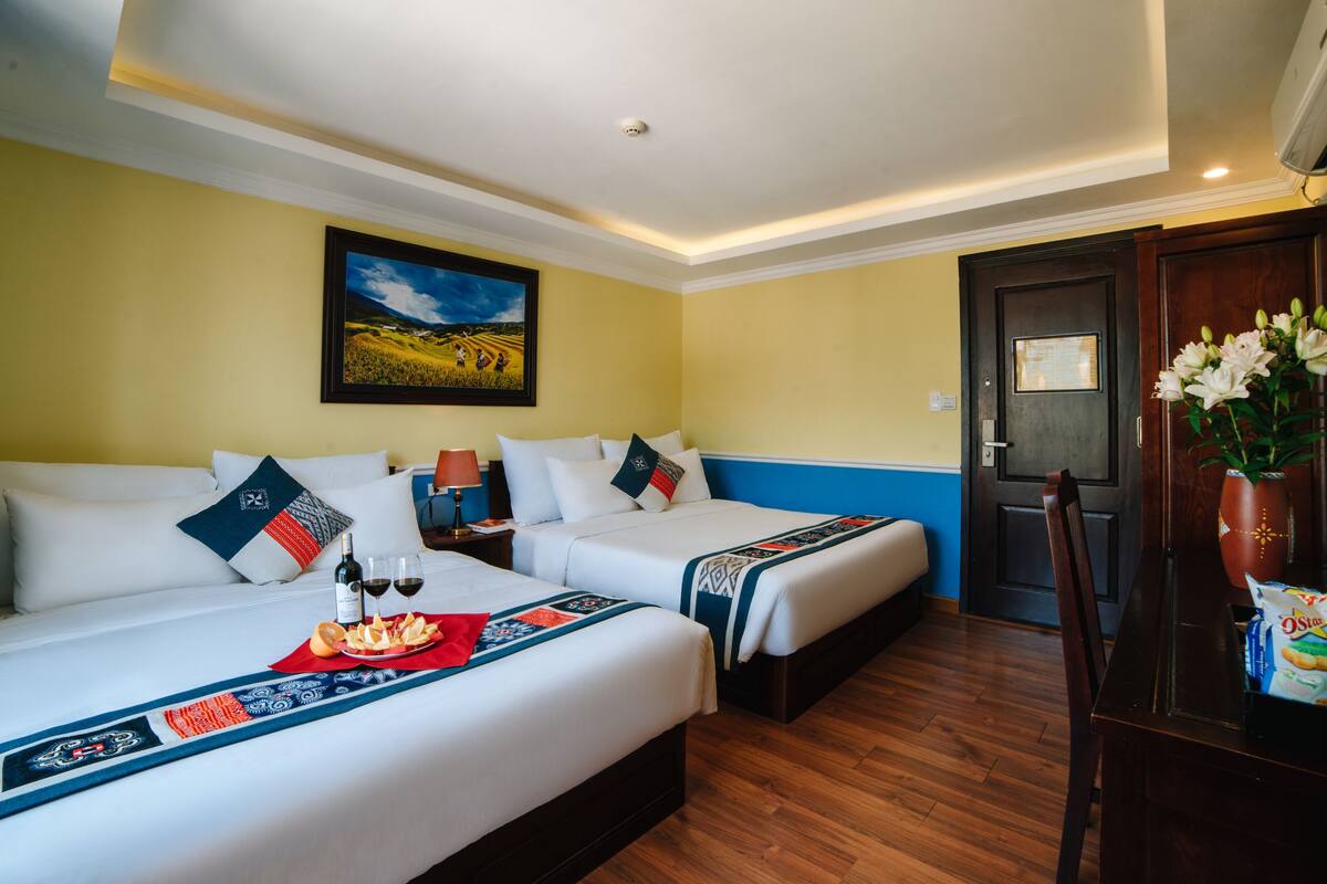 A hotel room with two beds, a wooden floor, a painting on the wall, and a door with a wooden frame.