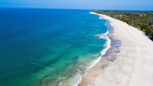 Vista aérea de una playa con aguas turquesas cristalinas, una orilla arenosa y una hilera de árboles y edificios a lo largo de la costa.